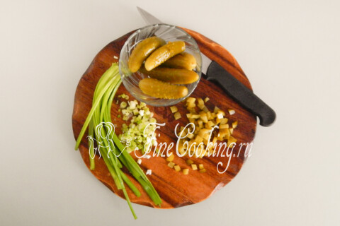 Маринованные огурчики и зелёный лук, мелко нарезанные на разделочной доске.